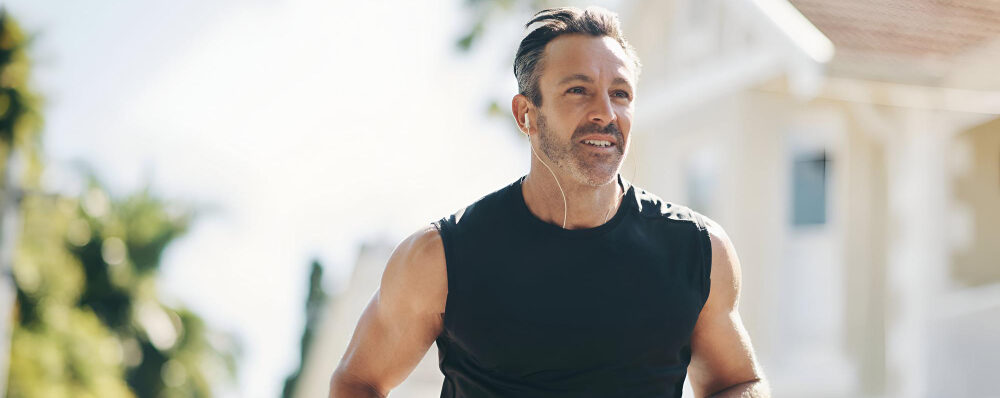 Homem de meia-idade correndo ao ar livre, usando camiseta preta sem mangas e fones de ouvido, com expressão determinada.