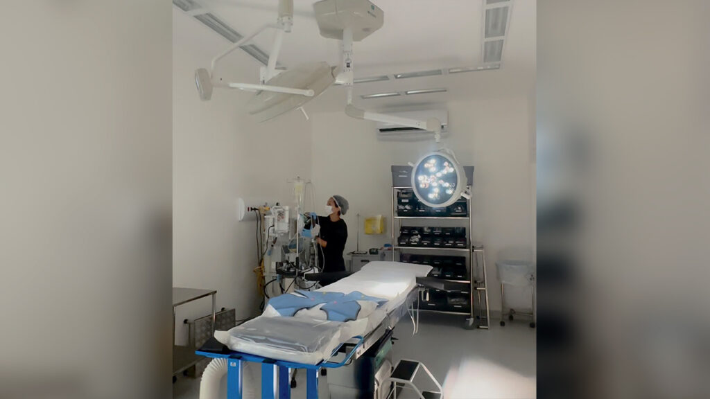 Profissional de saúde com máscara e touca organiza equipamentos ao lado de uma mesa cirúrgica em uma sala de cirurgia iluminada.