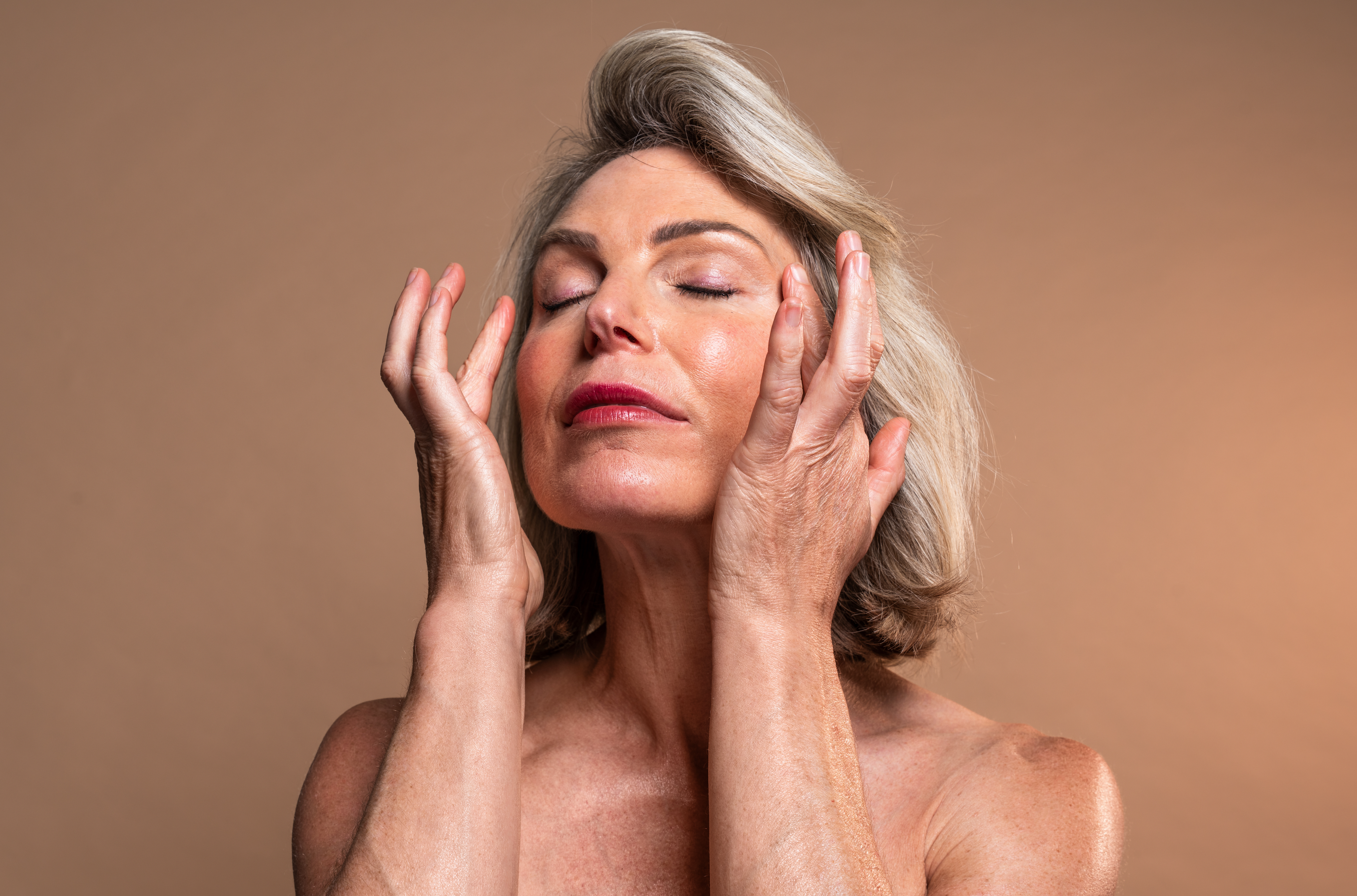 Mulher madura de cabelos grisalhos, olhos fechados, tocando o rosto com as mãos em gesto de cuidado com a pele, sobre fundo neutro.