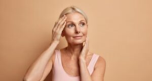 Mulher madura tocando o rosto e observando a pele com expressão serena.