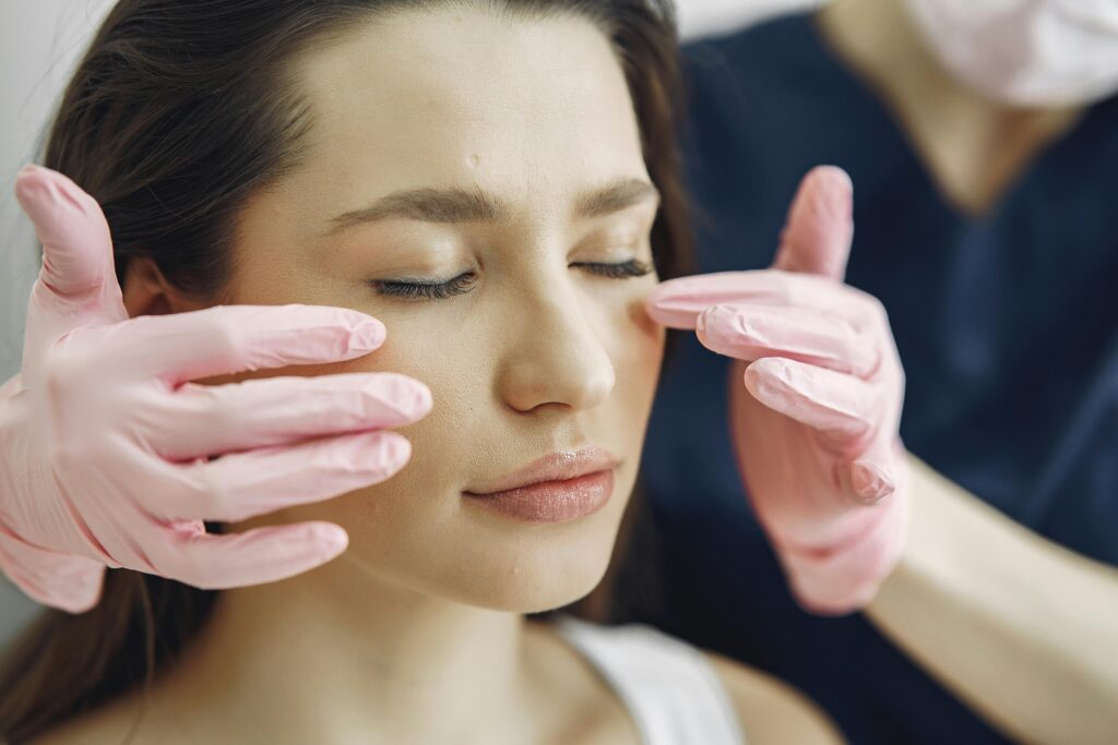 Profissional com luvas avaliando o rosto de mulher, focando na região das bochechas, com possível flacidez facial