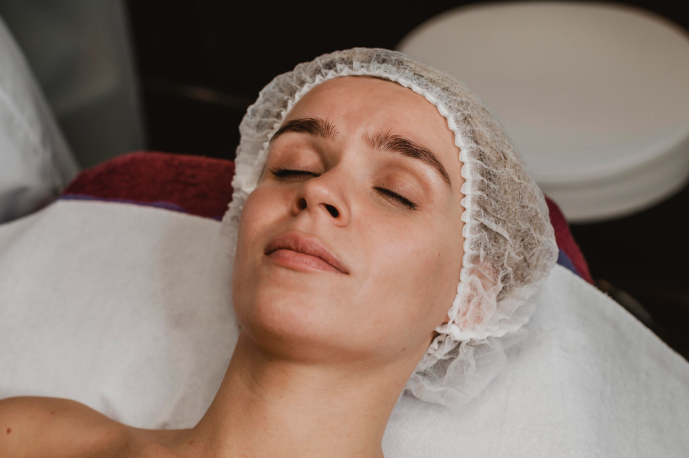 Mulher deitada em maca de clínica estética com os olhos fechados e touca descartável durante procedimento facial.