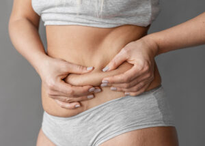 Mulher segurando a gordura abdominal com as mãos, destacando a região da barriga em roupa íntima cinza.