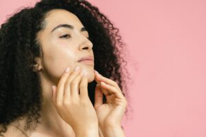 Mulher jovem com cabelos cacheados escuros tocando suavemente o queixo e a mandíbula com as pontas dos dedos, em frente a um fundo rosa sólido.