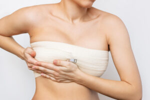 Foto em close-up do torso de uma mulher com uma bandagem compressiva elástica branca envolta firmemente no peito, área onde estão as próteses. Ela usa as duas mãos para segurar e ajustar a bandagem, contra um fundo branco e limpo.