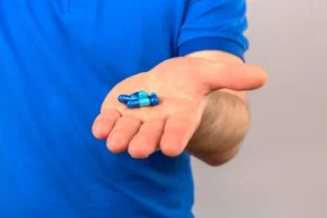 Pessoa com camiseta azul segurando algumas cápsulas de medicamentos azuis na palma da mão.