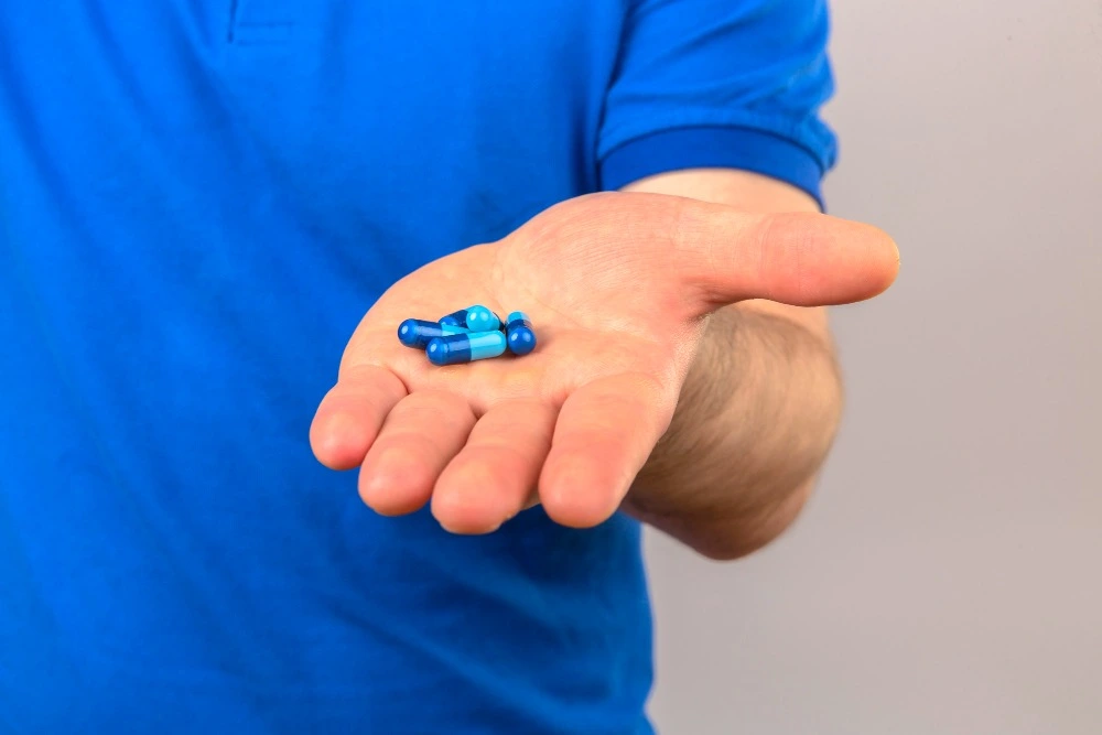 Pessoa com camiseta azul segurando algumas cápsulas de medicamentos azuis na palma da mão.