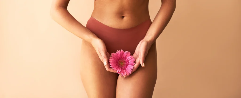 Fotografia do torso e coxas de uma mulher vestindo lingerie terracota contra um fundo bege. Ela segura delicadamente uma flor rosa vibrante com as duas mãos em frente à região pélvica, simbolizando o autocuidado e a saúde íntima feminina.