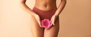 Fotografia do torso e coxas de uma mulher vestindo lingerie terracota contra um fundo bege. Ela segura delicadamente uma flor rosa vibrante com as duas mãos em frente à região pélvica, simbolizando o autocuidado e a saúde íntima feminina.